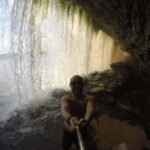 A selfie taken inside a cave behind a waterfall in Canaima, Venezuela, with water cascading down in the background.