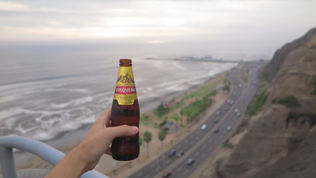 Best Places to Visit in Lima - A hand holding a cold bottle of Cusqueña beer at Larcomar Shopping Centre in Lima, Peru, with the dramatic Pacific Ocean coastline, cliffs, highway, and a misty sky visible in the background.