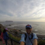 Best Places to Visit in Lima - A traveler with a cap and glasses taking a selfie on the Miraflores cliffs near Parque del Amor, overlooking the Pacific Ocean in Lima, Peru.