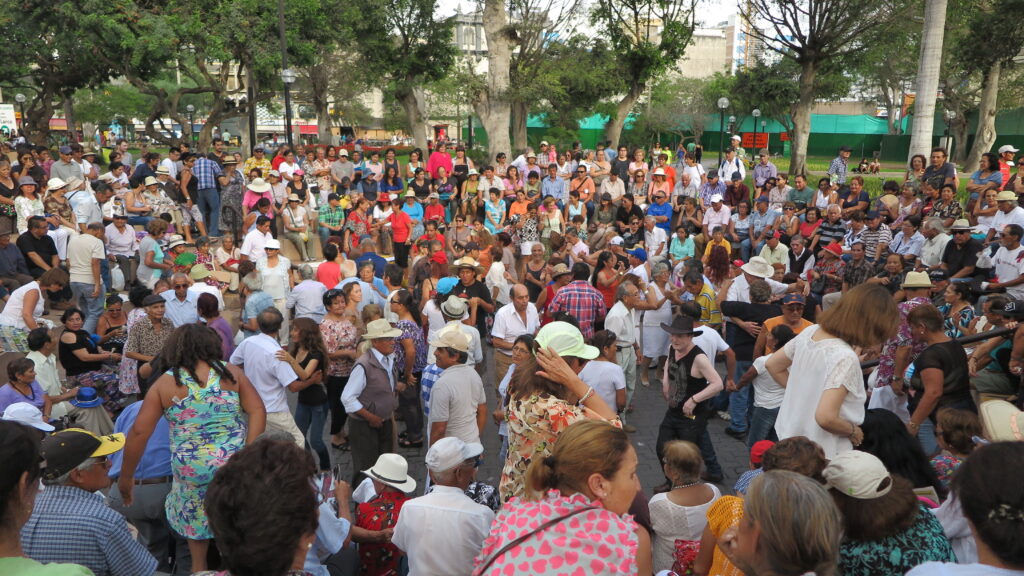 Best Places to Visit in Lima - A large crowd of people dancing outdoors in Parque 7 de Junio, Lima. Couples are dancing in the center, surrounded by hundreds of spectators and trees in the background.