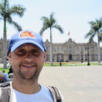 Best Places to Visit in Lima - A selfie of a smiling man at the historic Plaza Mayor (Plaza de Armas) in Lima, Peru. He wears a blue baseball cap and a white t-shirt. The Government Palace of Peru (Palacio de Gobierno), with the Peruvian flag flying, is prominent in the background, surrounded by palm trees and other historic buildings under a sunny sky.