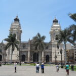 Best Places to Visit in Lima - A wide-angle photograph of the historic Plaza Mayor (Plaza de Armas) in Lima, Peru. The grand Cathedral of Lima (Catedral de Lima), with its two tall bell towers, is central to the image. To its left is the Archbishop's Palace, featuring traditional enclosed wooden balconies. The square is adorned with palm trees and has pedestrians walking. Yellow colonial buildings are on the right. The sky is clear blue.