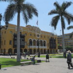 The yellow Municipal Palace of Lima (Palacio Municipal de Lima) located at Plaza Mayor in Lima, Peru. The building features dark wooden balconies, arches, palm trees, and street lamps in the foreground.