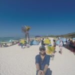 A man takes a selfie on a white-sand beach with colorful tents, people relaxing, and a clear blue sky at La Tortuga Island, Venezuela.