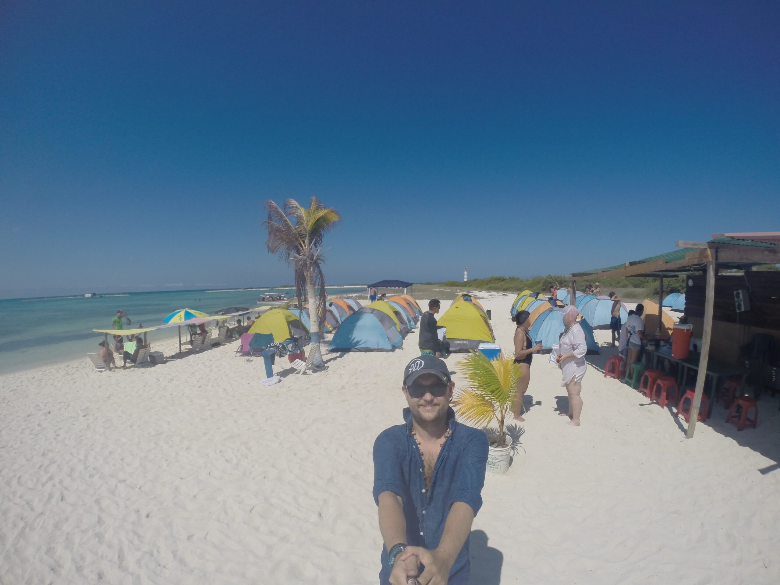 A man takes a selfie on a white-sand beach with colorful tents, people relaxing, and a clear blue sky at La Tortuga Island, Venezuela.