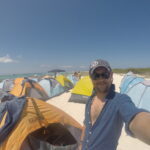 A man takes a selfie among rows of colorful camping tents on the beach at La Tortuga Island under a clear blue sky.