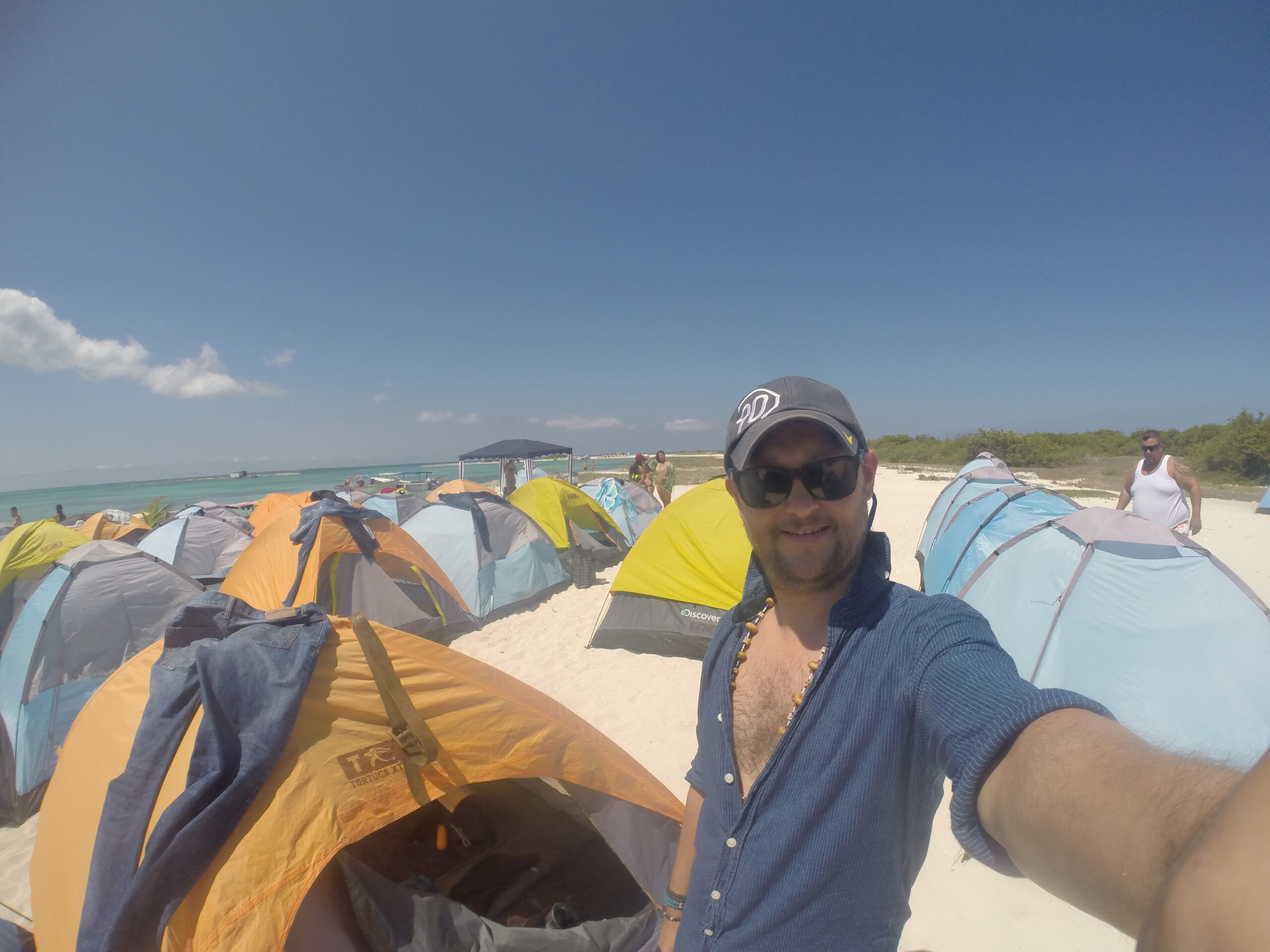 A man takes a selfie among rows of colorful camping tents on the beach at La Tortuga Island under a clear blue sky.