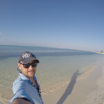 Selfie of a male traveler smiling on a white-sand beach with turquoise water and a clear blue sky on La Tortuga Island, Venezuela.