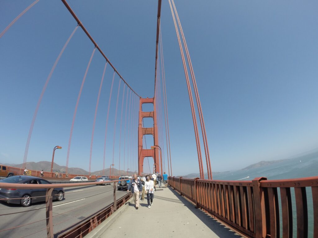 Golden Gate Köprüsü'nün yaya yolunda çekilmiş geniş açılı fotoğraf; yürüyen insanlar, bitişikteki yolda ilerleyen araçlar ve turuncu kuleye doğru uzanan asma kabloları görülüyor.