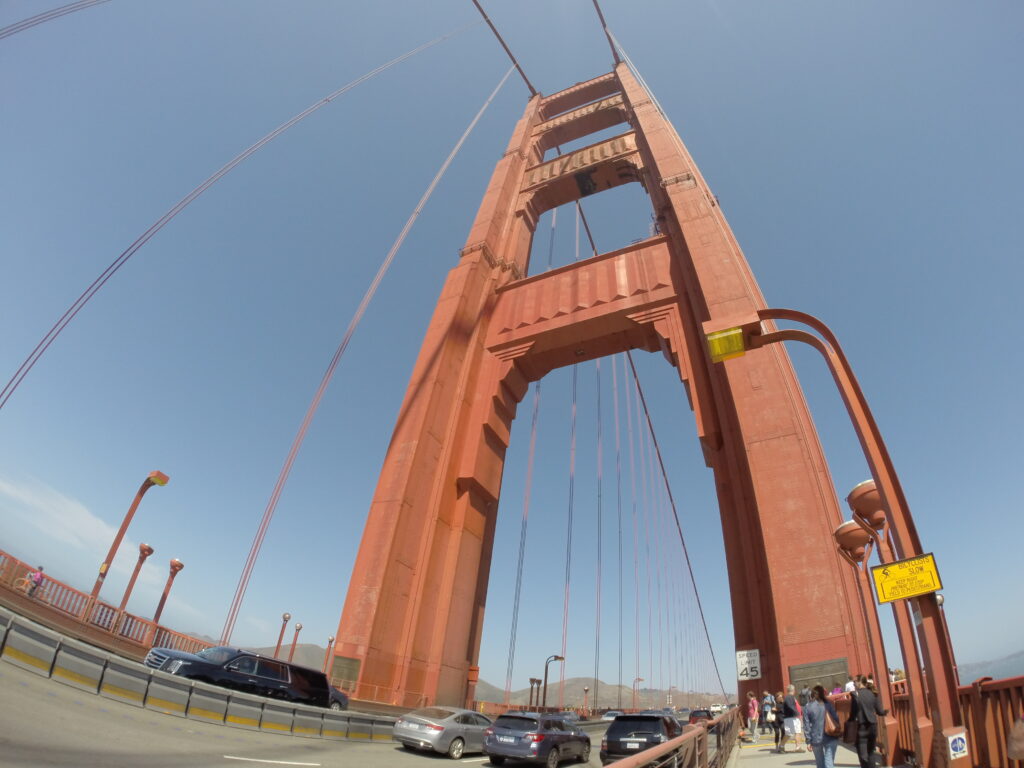 Golden Gate Köprüsü'nün yüksek turuncu kulesine aşağıdan yukarıya doğru çekilmiş geniş açılı bakış; yolda ilerleyen araçlar ve kaldırımda yürüyen yayalar görülüyor.