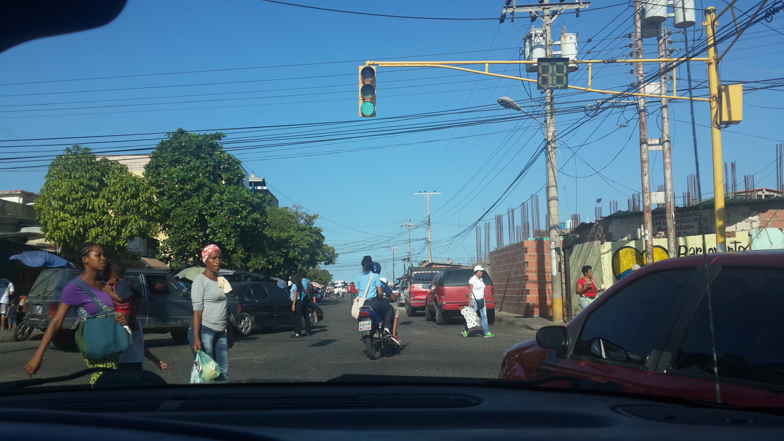 Street scene in Higuerote, Venezuela, showing people walking, a motorbike passing, and cars under a clear blue sky.