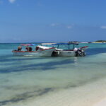 Two fishing boats anchored near the shore of La Tortuga Island with beach tents and clear blue skies in the background.