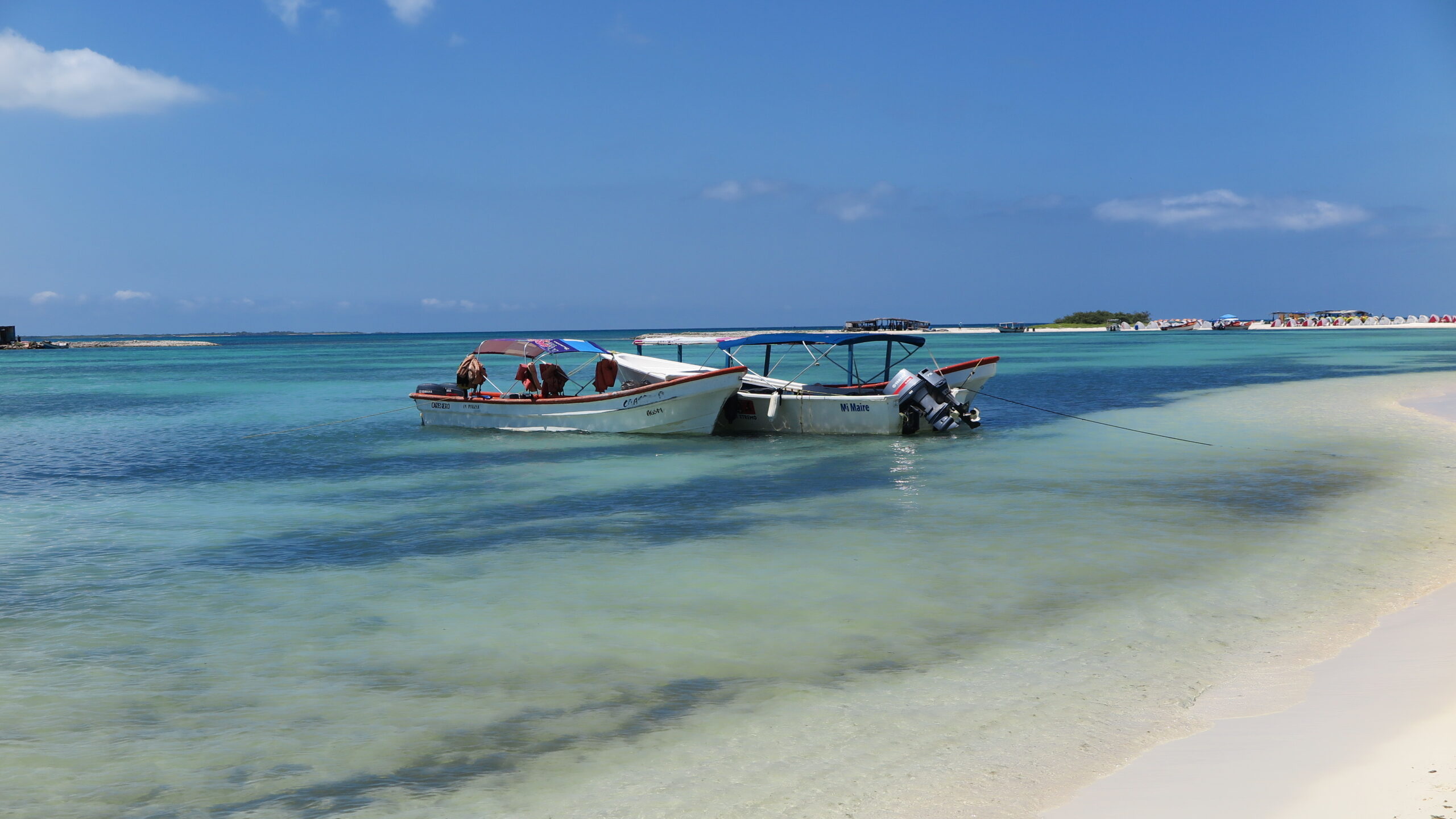 Two fishing boats anchored near the shore of La Tortuga Island with beach tents and clear blue skies in the background.