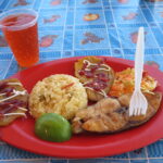 A red plate with grilled fish, salad, rice, potatoes, and lime slices, with a red drink in a plastic cup behind it. Photo taken on La Tortuga Island, Venezuela.