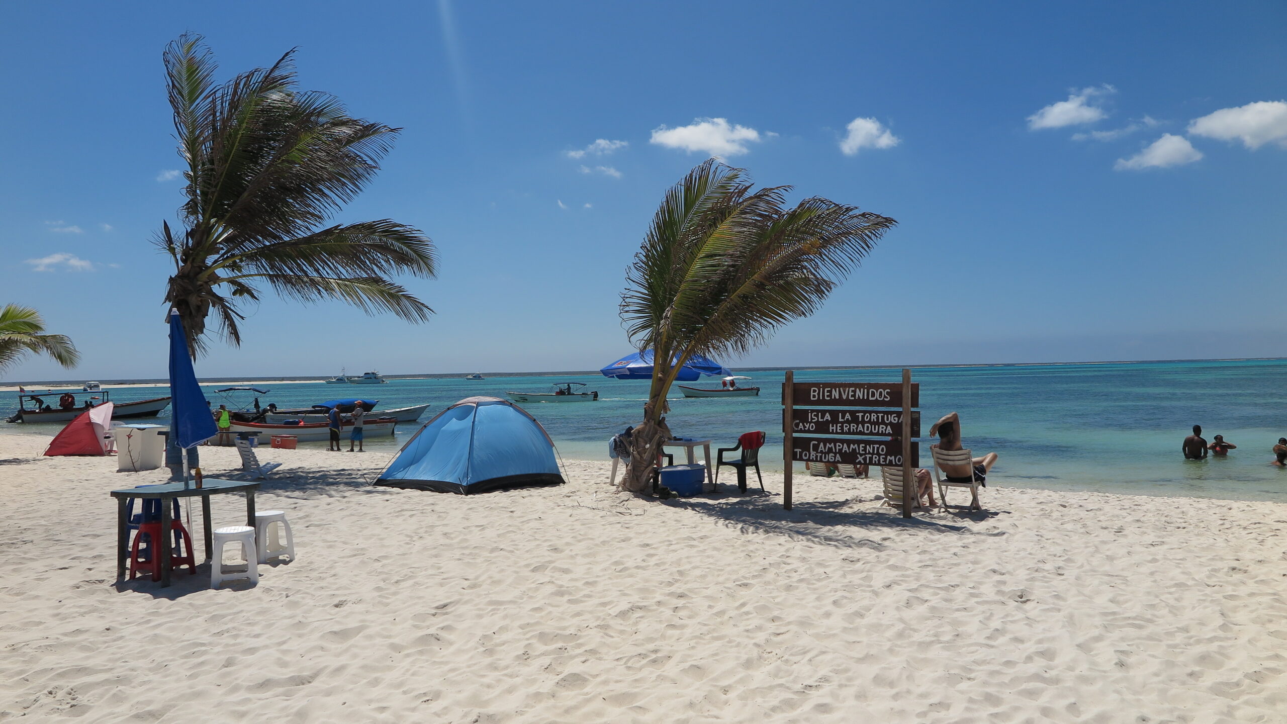 Beach tents and palm trees along the shore of La Tortuga Island, with people swimming and boats anchored in turquoise waters.