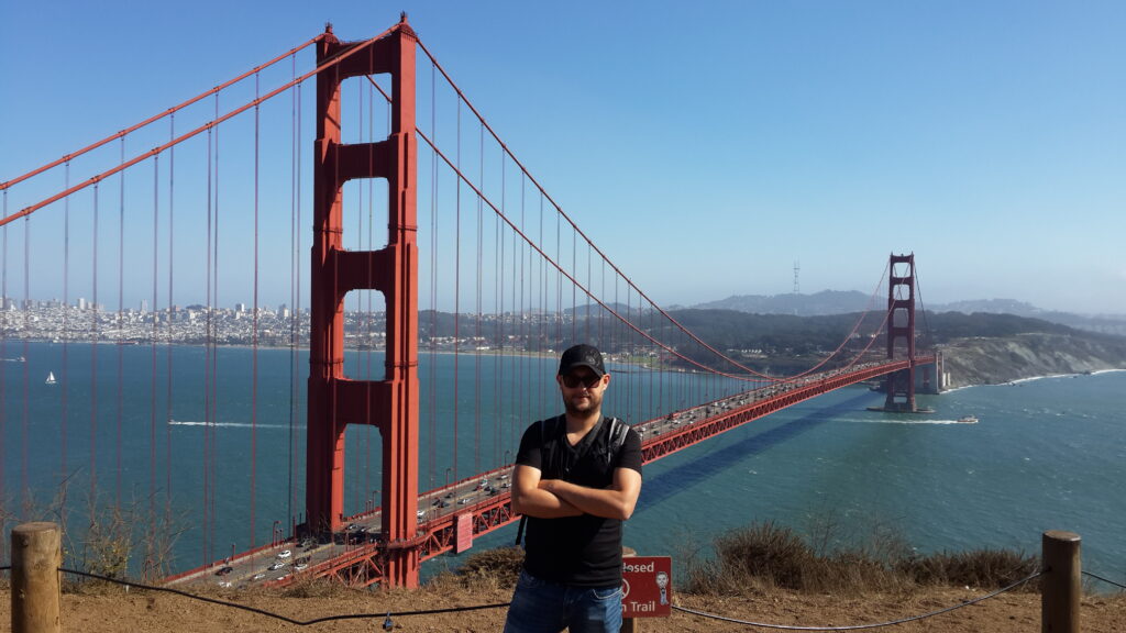 Battery Spencer seyir noktasında kollarını kavuşturarak duran; siyah tişörtlü, şapkalı ve güneş gözlüklü bir adam; arka planda panoramik Golden Gate Köprüsü ve San Francisco şehir manzarası.