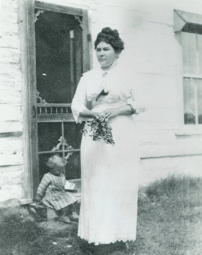 Black and white photo of Annie Huble standing in front of an old building, with a small child seated nearby.