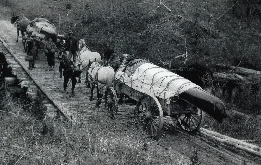 Black and white photo of people crossing a wooden bridge at Giscome Portage with horse-drawn carts carrying a wooden boat and goods.