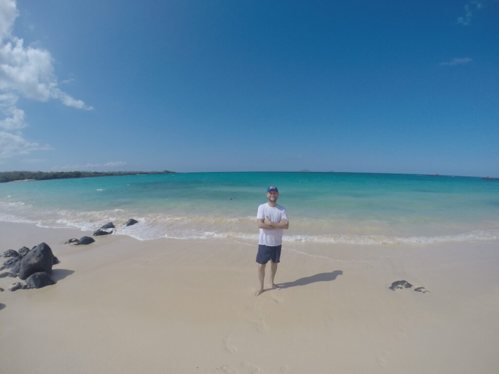 A person standing on a yellow sandy beach in the Galapagos Islands, with turquoise water and small rocks around, under a sunny sky.