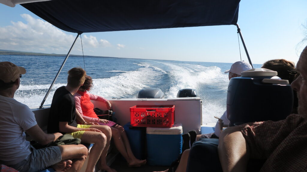 People seated on a boat, facing the white wake trailing behind as the vessel sails north through calm blue waters under clear skies in the Galapagos Islands.