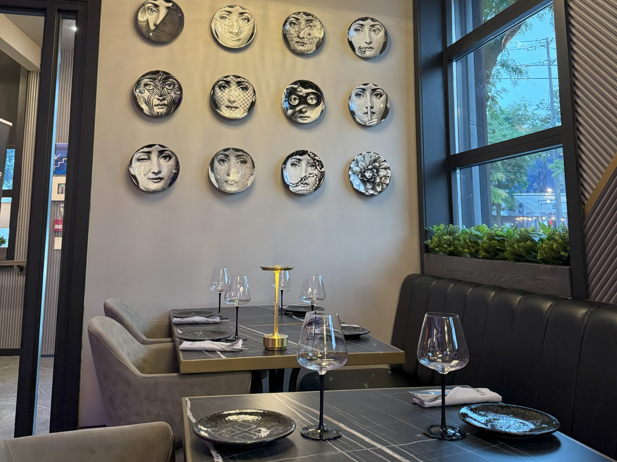 Elegant restaurant seating area with wall-mounted decorative plates featuring artistic faces, black leather banquette, and neatly set tables.