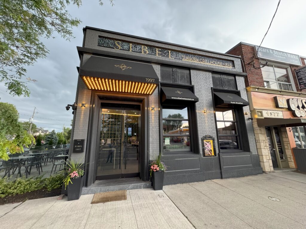 A view of the entrance to Sibel Restaurant, featuring a modern glass facade, warm lighting, and the restaurant's name displayed above the door.