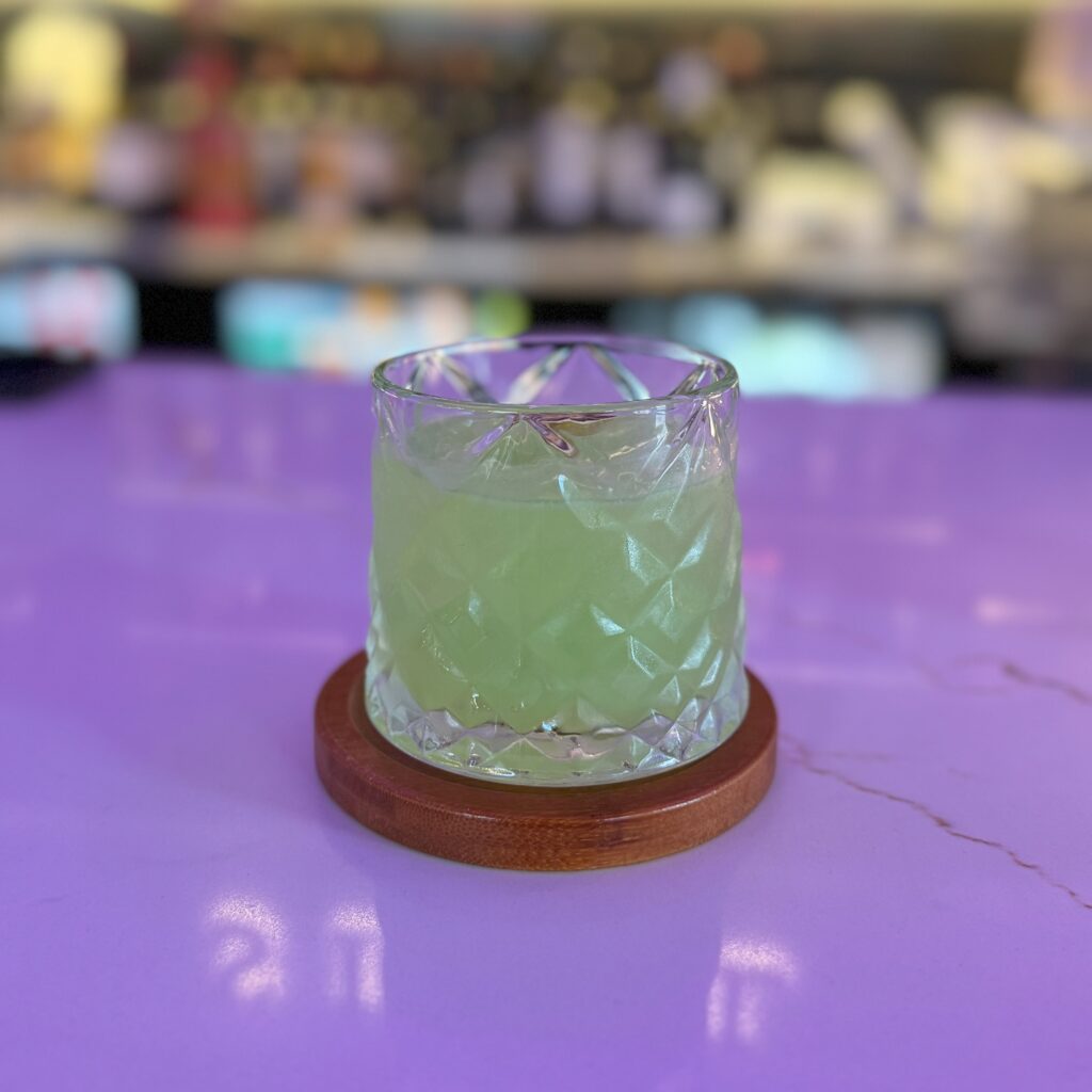 The Sultan’s Special cocktail, a vibrant green drink served in a whiskey-style glass on the bar counter, with the bar softly blurred in the background.