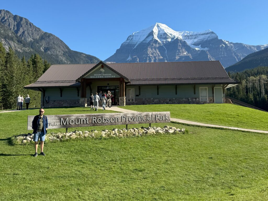 Traveler standing in front of the Mount Robson Provincial Park sign with the Welcome Centre and Mount Robson summit in the background.