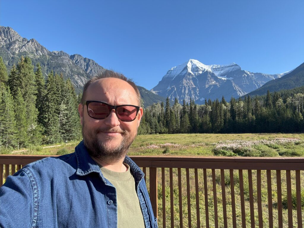 A man takes a selfie on the porch behind the Mount Robson Welcome Centre with Mount Robson’s snow-capped peak rising in the background.