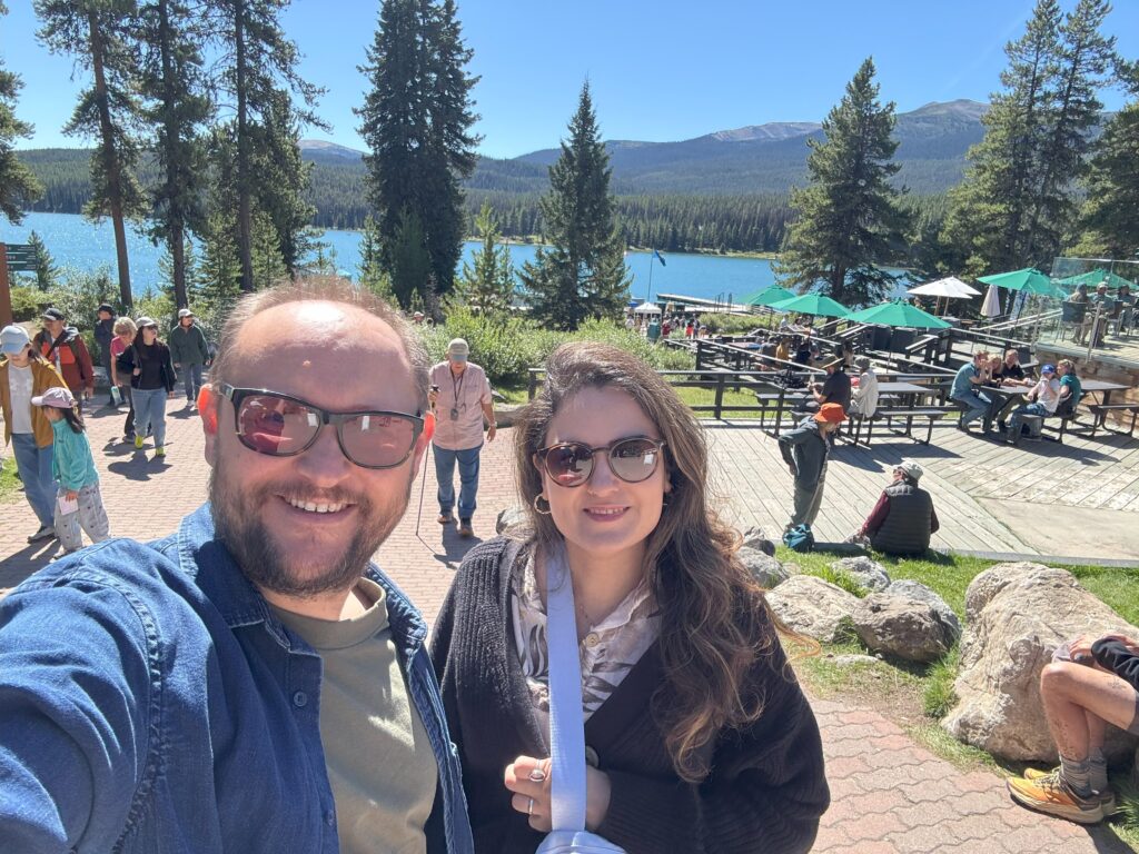 A couple takes a selfie at Maligne Lake with crowds of visitors and turquoise waters in the background.