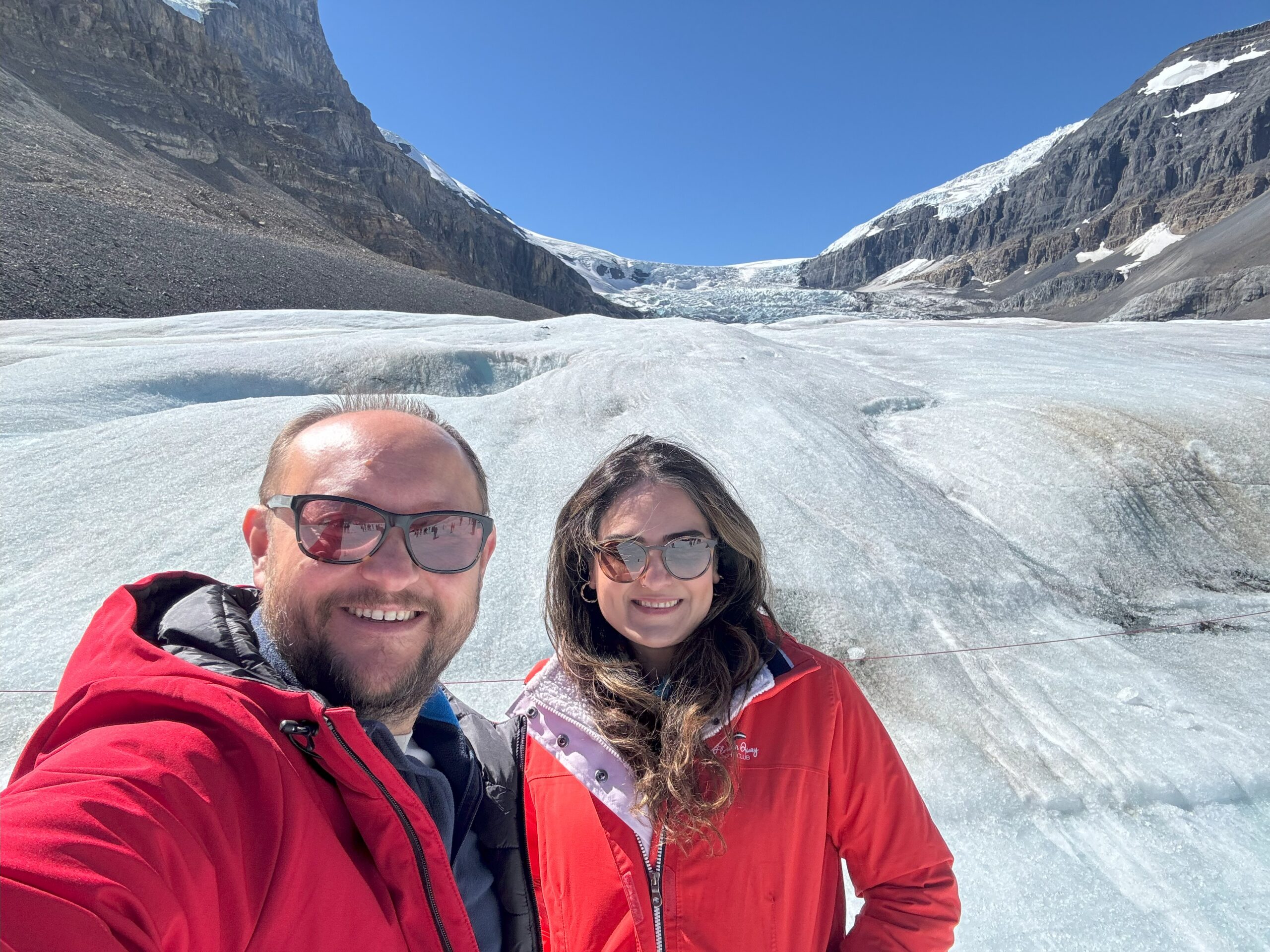 A couple smiles for a selfie on the Athabasca Glacier in Jasper National Park, surrounded by ice, rugged mountain slopes, and a clear blue sky.