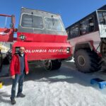 Bir adam, açık mavi gökyüzünün altında Athabasca Buzulu'nda "Columbia Icefield" etiketli büyük kırmızı bir Ice Explorer aracının yanında duruyor.