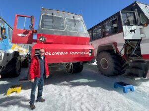 Bir adam, açık mavi gökyüzünün altında Athabasca Buzulu'nda "Columbia Icefield" etiketli büyük kırmızı bir Ice Explorer aracının yanında duruyor.