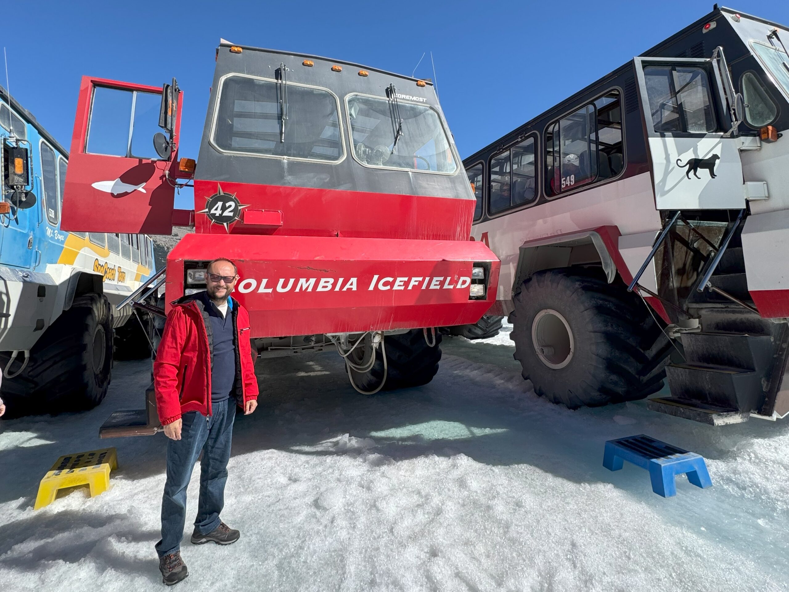 A man stands beside a large red Ice Explorer vehicle labeled “Columbia Icefield” on the Athabasca Glacier under a clear blue sky.