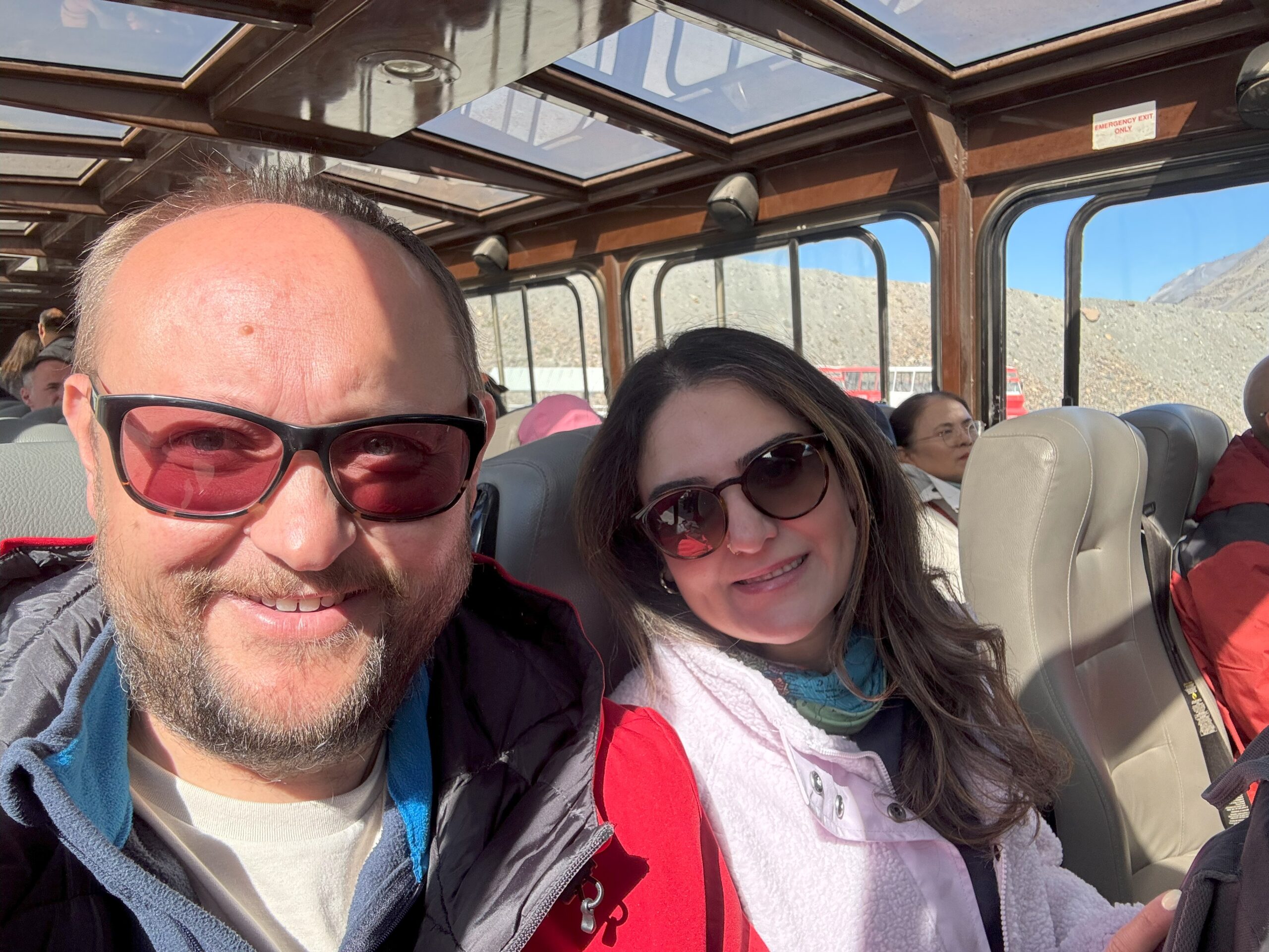 A couple smiles inside the Ice Explorer vehicle during the Athabasca Glacier tour, with sunlight streaming through the glass roof and other passengers seated behind them.