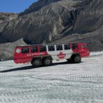 Üzerinde "Kanada" yazan büyük, kırmızı ve beyaz bir Ice Explorer aracı, kayalık dağlık araziyle çevrili Athabasca Buzulu'nun buzlu yüzeyinde ilerliyor.