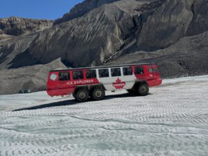 Üzerinde "Kanada" yazan büyük, kırmızı ve beyaz bir Ice Explorer aracı, kayalık dağlık araziyle çevrili Athabasca Buzulu'nun buzlu yüzeyinde ilerliyor.