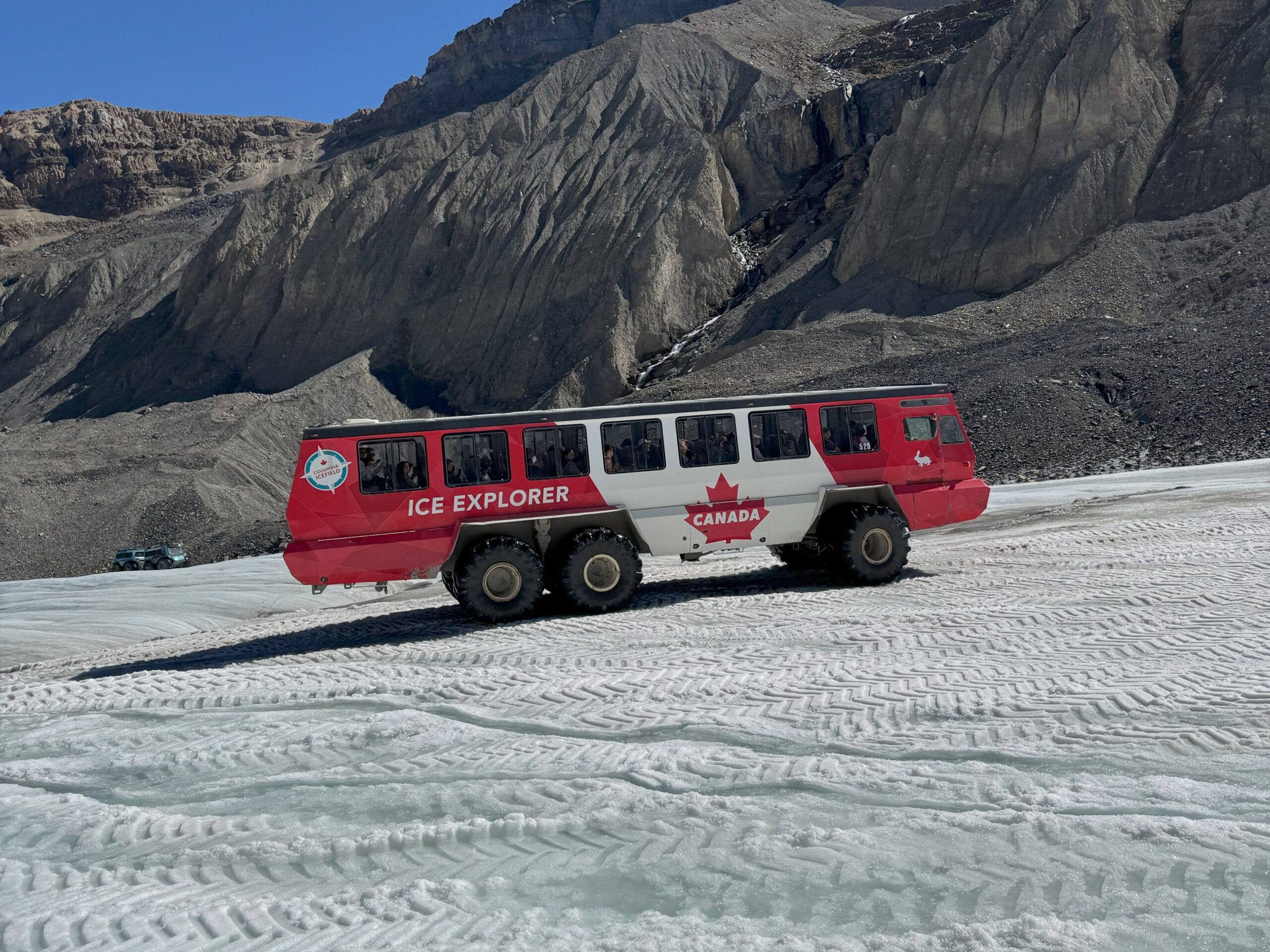 A large red and white Ice Explorer vehicle labeled "Canada" drives across the icy surface of Athabasca Glacier, surrounded by rocky mountain terrain.