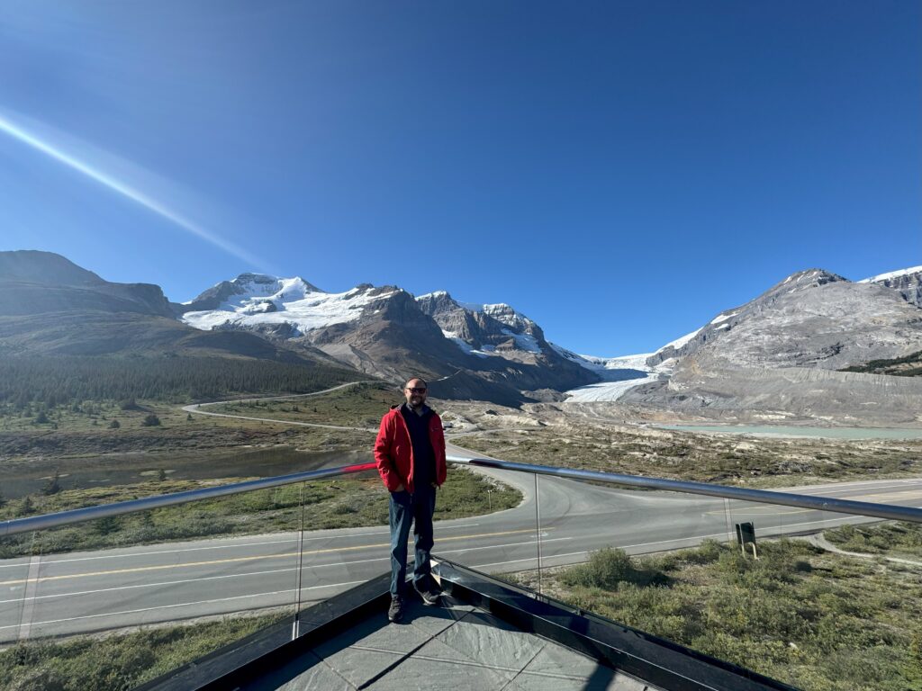 Kırmızı ceketli bir kişi parlak mavi gökyüzünün altında Jasper Milli Parkı'ndaki Athabasca Buzulu'na bakan Icefield Information Centre'nin terasında duruyor.