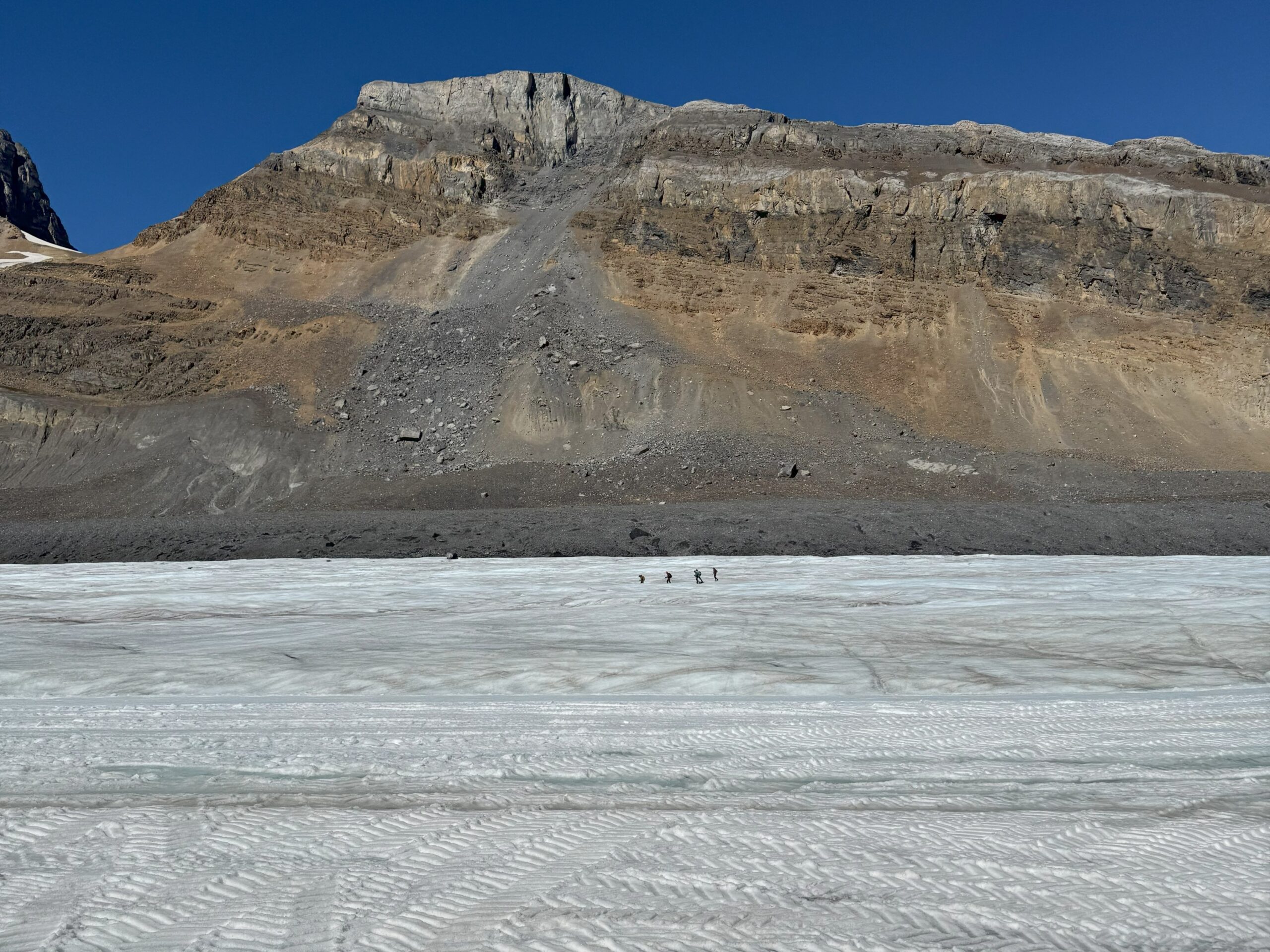 View of the rocky road leading to Athabasca Glacier with people walking on the ice in the distance.
