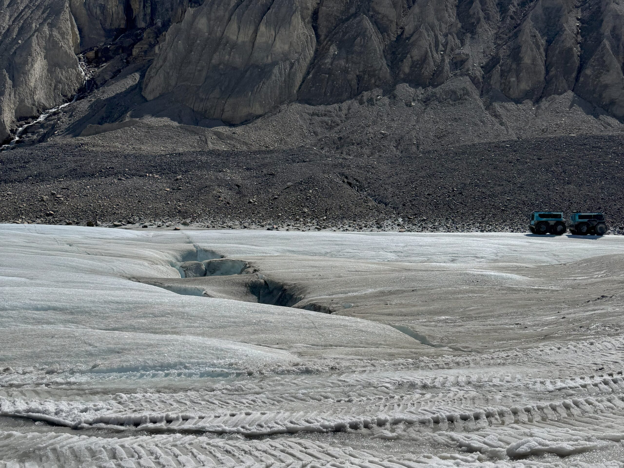 View of Athabasca Glacier with visible crevasses, rocky mountain slopes, and a glacier tour vehicle on the ice and road.