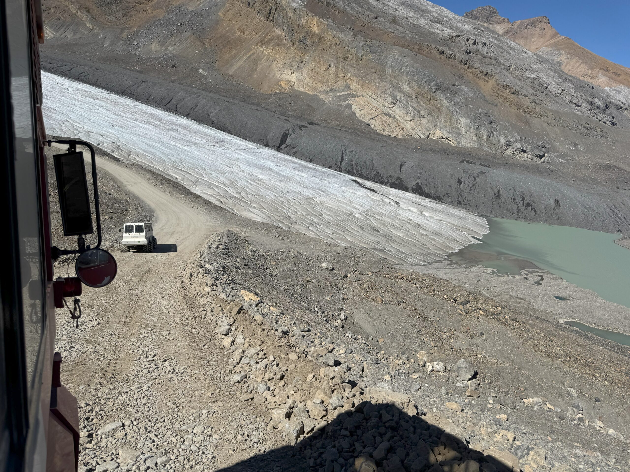 View from an Ice Explorer vehicle descending a steep, rocky road toward Athabasca Glacier, with another glacier vehicle ahead and a meltwater lake visible beside the ice.