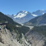 Açık mavi gökyüzünün altında, arka planda karla kaplı Rocky Dağları ve buzulların bulunduğu derin ormanlık Sunwapta vadisinin üzerinde uzanan Columbia Icefield Skywalk manzarası.