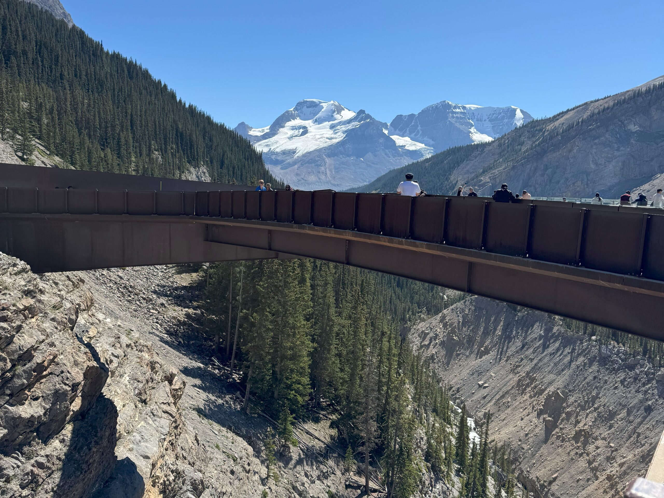Visitors standing on the Columbia Icefield Skywalk, a glass-floored platform extending over a deep valley, with forested slopes and snow-covered mountains in the background.
