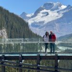 Bir çift, arka planda karla kaplı dağ zirveleri ve buzul vadisiyle birlikte cam tabanlı Columbia Icefield Skywalk üzerinde duruyor.