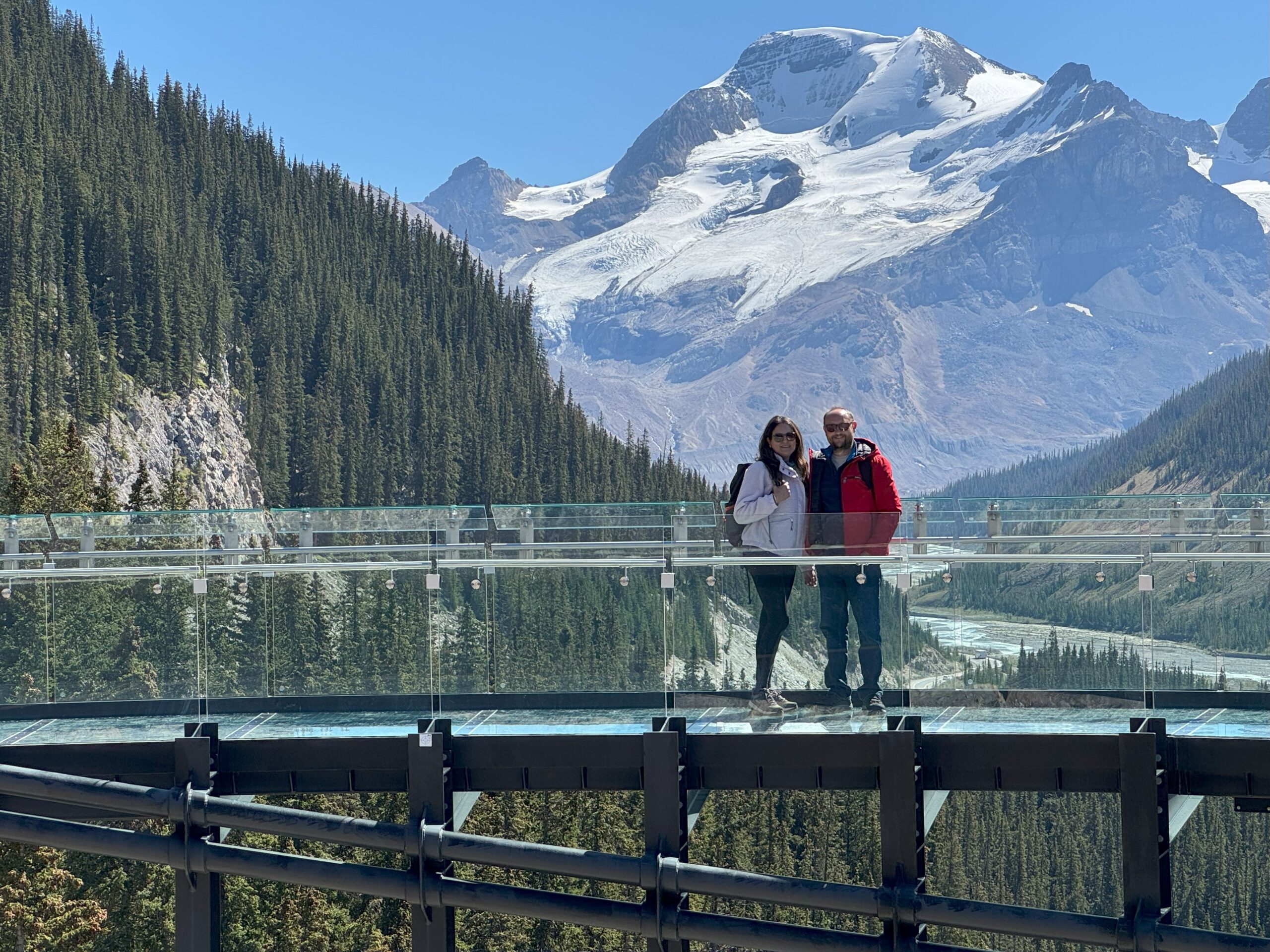 A couple standing on the glass-floored Columbia Icefield Skywalk with towering snow-covered peaks and a glacier valley in the background.