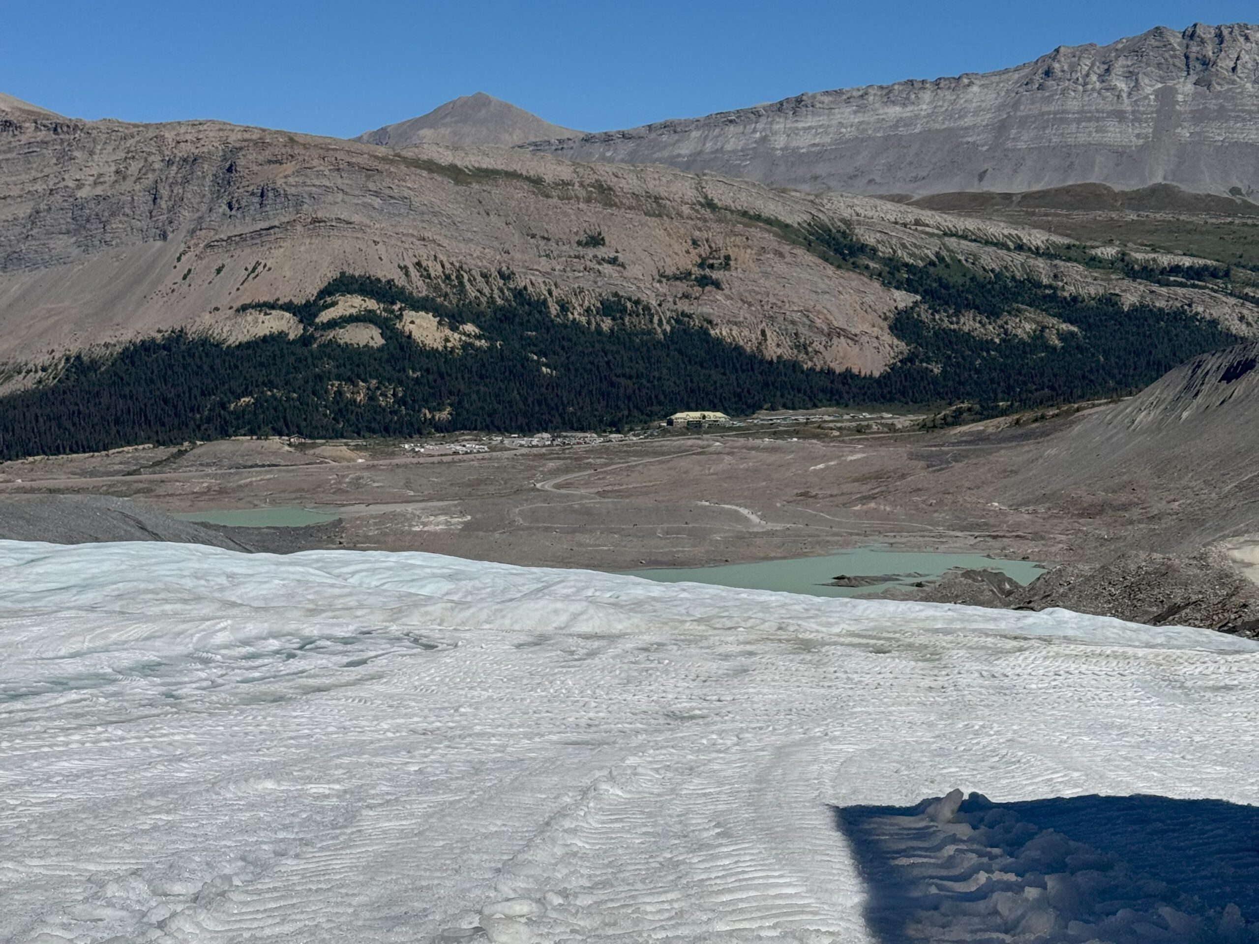 View from the Athabasca Glacier showing icy terrain in the foreground, with rocky mountains, green valleys, and glacial meltwater ponds below under a clear blue sky.