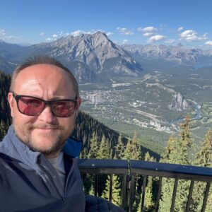 A person takes a selfie on the Sulphur Mountain observation deck with a panoramic view of Banff town and surrounding Rocky Mountains in the background.
