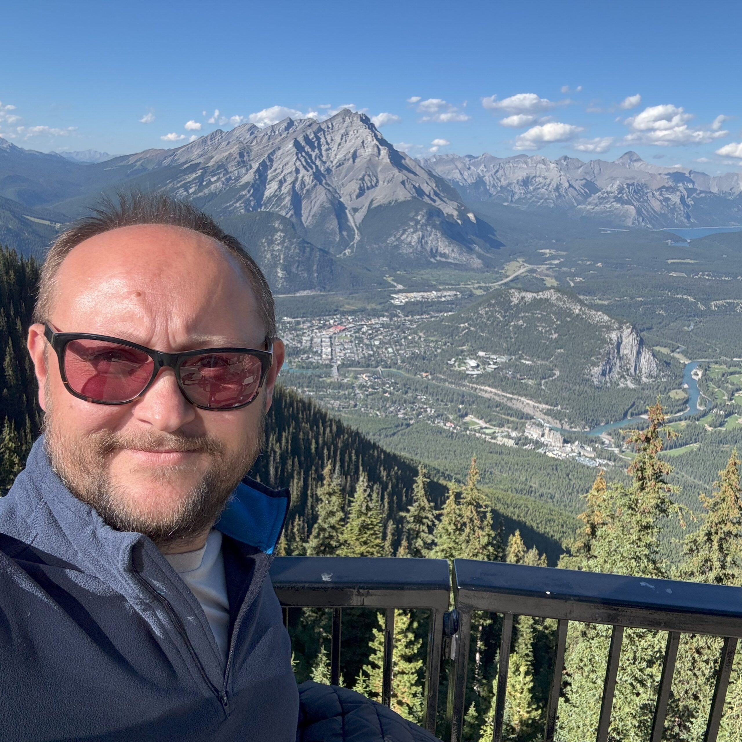 A person takes a selfie on the Sulphur Mountain observation deck with a panoramic view of Banff town and surrounding Rocky Mountains in the background.