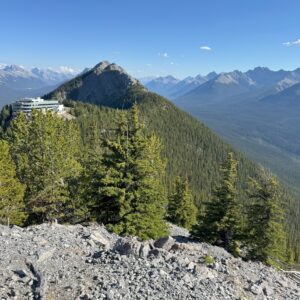 View from Sulphur Mountain in Banff, showing the Banff Gondola station on the left and a forested mountain ridge stretching into the distance under a clear blue sky.